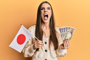 Beautiful brunette young woman holding japan flag and yen banknotes angry and mad screaming...