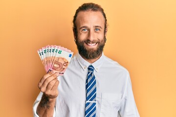 Handsome business man with beard and long hair holding bunch of 10 euro banknotes looking positive and happy standing and smiling with a confident smile showing teeth