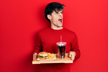 Handsome hipster young man eating a tasty classic burger with fries and soda angry and mad screaming frustrated and furious, shouting with anger. rage and aggressive concept.
