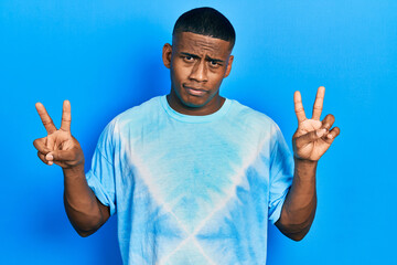 Young black man doing victory sign and peace gesture skeptic and nervous, frowning upset because of...
