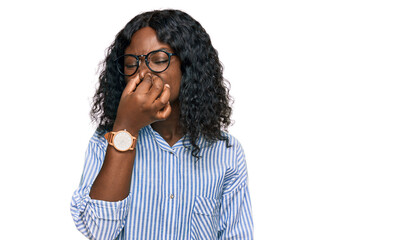 Fototapeta na wymiar Beautiful african young woman wearing casual clothes and glasses smelling something stinky and disgusting, intolerable smell, holding breath with fingers on nose. bad smell