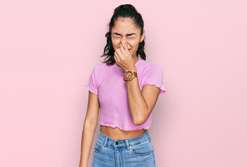 Hispanic teenager girl with dental braces wearing casual clothes smelling something stinky and...