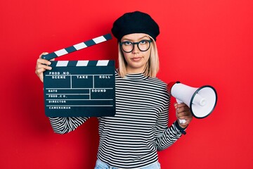 Beautiful blonde woman holding video film clapboard and megaphone skeptic and nervous, frowning...