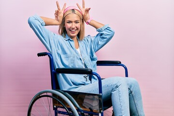Beautiful blonde woman sitting on wheelchair posing funny and crazy with fingers on head as bunny...