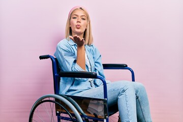Beautiful blonde woman sitting on wheelchair looking at the camera blowing a kiss with hand on air...