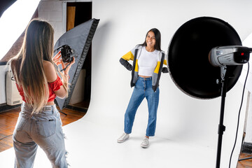Photo session of a model and a woman photographer in a photography studio with flashes and a white background. Fashion concept. - obrazy, fototapety, plakaty