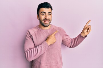 Young hispanic man wearing casual clothes smiling and looking at the camera pointing with two hands and fingers to the side.