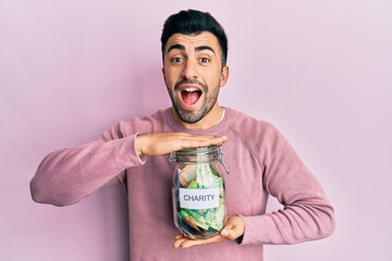 Young hispanic man holding charity jar with money celebrating crazy and amazed for success with open eyes screaming excited.