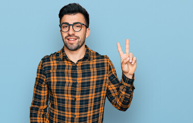 Naklejka premium Hispanic man with beard wearing casual clothes and glasses smiling with happy face winking at the camera doing victory sign. number two.
