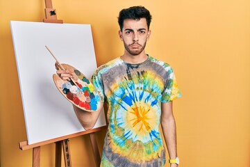 Young hispanic man standing drawing with palette by painter easel stand depressed and worry for...