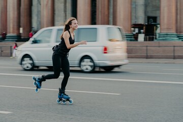 Outdoor shot of happy smiling woman spends time in active way rides on skating rollers demonstrated high speed during motion poses on roaad with transport in background. Hobby and lifestyle.