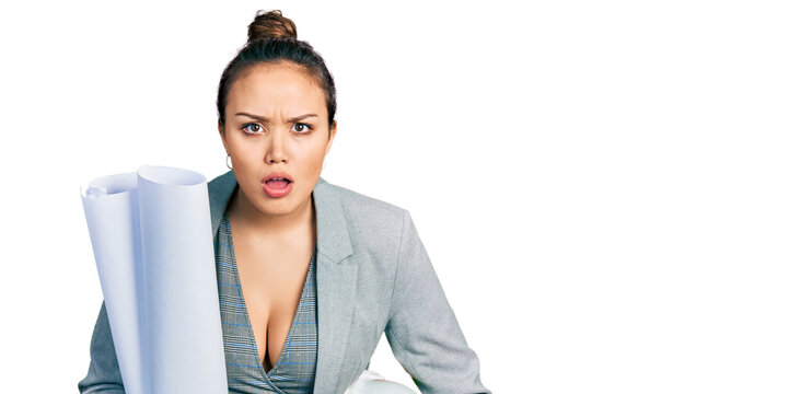 Young hispanic girl holding safety helmet and blueprints afraid and shocked with surprise and amazed expression, fear and excited face.