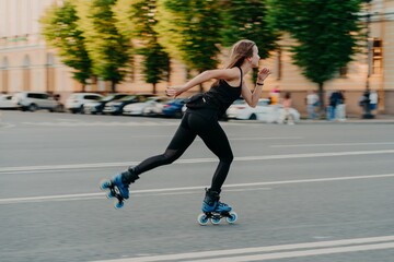 Professional female roller demonstrates her abilities of rollerblading rides very quckly on road along city enjoys sunny day dressed in black active wear. Active lifestyle hobby and fitness activity - obrazy, fototapety, plakaty
