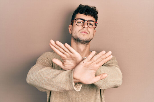 Young hispanic man wearing casual clothes and glasses rejection expression crossing arms and palms doing negative sign, angry face