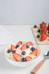 Quark fresh dairy product made of fermented soured milk also curd or cottage cheese served in bowl with spoon and fresh strawberries and blueberries on cutting board on white wooden table. Vertical