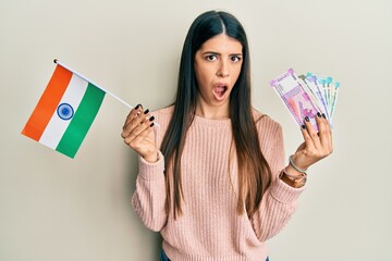 Young hispanic woman holding india flag and rupee banknotes in shock face, looking skeptical and sarcastic, surprised with open mouth