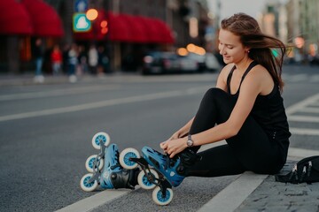 Outdoor activities fit lifestyle. Sideways shot of active woman sits on road adjustes rollerblades...