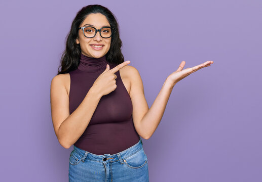 Brunette young woman wearing casual clothes and glasses amazed and smiling to the camera while presenting with hand and pointing with finger.