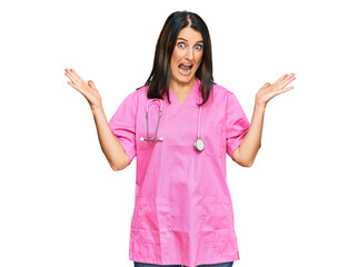 Middle age brunette woman wearing doctor uniform and stethoscope celebrating victory with happy smile and winner expression with raised hands