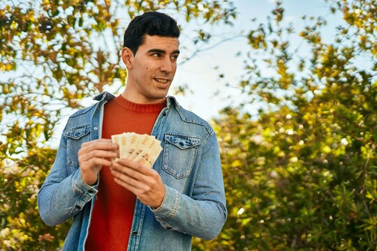 Young hispanic man smiling happy holding denmark krone banknotes at the city.