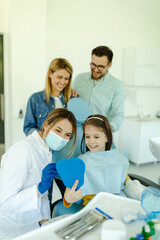 Girl looking in the mirror after dental procedure while parents standing near her for support.