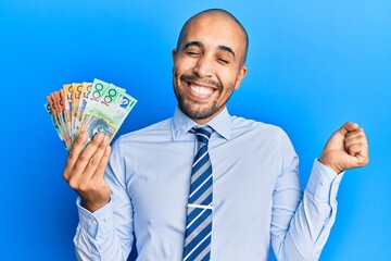 Hispanic adult man holding australian dollars screaming proud, celebrating victory and success very excited with raised arm