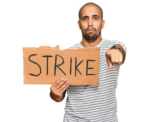 Hispanic adult man holding strike banner cardboard pointing with finger to the camera and to you, confident gesture looking serious