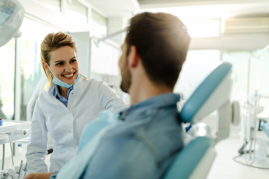 Beautiful Female Dentist Talking With Happy Male Patient At Clinic.