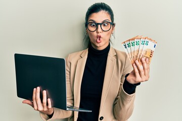 Young hispanic woman wearing business style holding laptop and 50 euros making fish face with mouth...