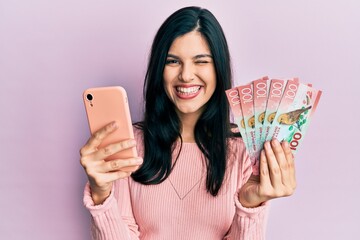Young hispanic woman using smartphone holding new zealand dollars banknotes winking looking at the...