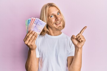 Caucasian young man with long hair holding thai baht banknotes smiling happy pointing with hand and finger to the side