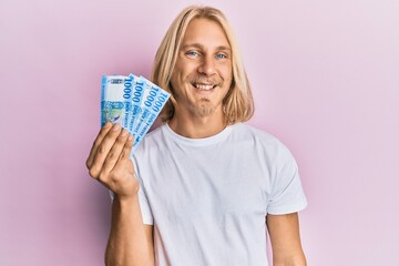 Caucasian young man with long hair holding 1000 hungarian forint banknotes looking positive and happy standing and smiling with a confident smile showing teeth