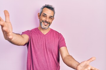 Handsome middle age man with grey hair wearing casual pink t shirt looking at the camera smiling with open arms for hug. cheerful expression embracing happiness.