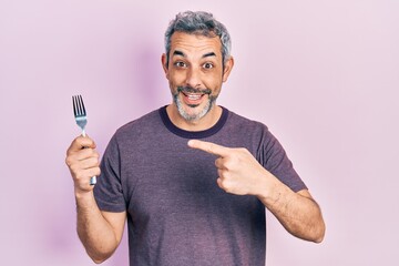 Handsome middle age man with grey hair holding one silver fork smiling happy pointing with hand and finger