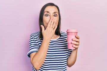 Middle age hispanic woman holding coffee covering mouth with hand, shocked and afraid for mistake....