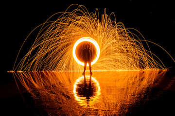 person posing standing on the beach with ring of fire behind. Reflections in the water. Steel wool lightpainting - Powered by Adobe