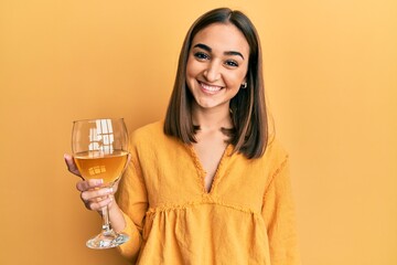 Young brunette girl drinking a glass of white wine looking positive and happy standing and smiling...