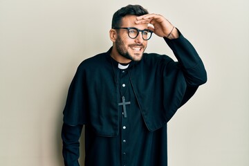 Young hispanic man wearing priest uniform standing over white background very happy and smiling looking far away with hand over head. searching concept.