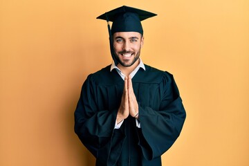 Young hispanic man wearing graduation cap and ceremony robe praying with hands together asking for...