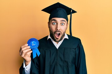 Young hispanic man wearing graduation robe holding 1 st place badge scared and amazed with open...