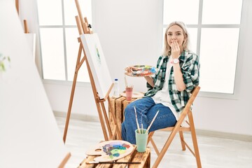 Young artist woman painting on canvas at art studio laughing and embarrassed giggle covering mouth...