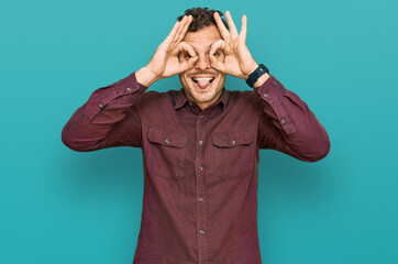 Young hispanic man wearing casual clothes doing ok gesture like binoculars sticking tongue out, eyes looking through fingers. crazy expression.