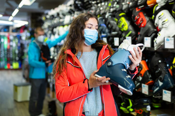 Positive female buyer in a medical protective face mask chooses ski boots in a sports equipment store