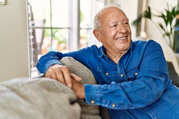 Fototapeta premium Senior man with grey hair sitting on the sofa at the living room of his house. Mature man smiling happy at home.