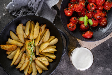 Roasted potatoes with rosemary in iron casserole and plate of confit tomatoes on dark background