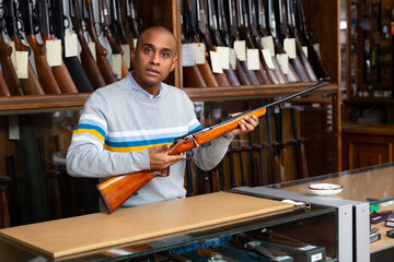 Hispanic salesman working behind counter of gun shop, showing hunting single-barrel rifle.