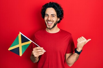 Handsome hispanic man holding jamaica flag pointing thumb up to the side smiling happy with open mouth