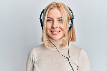 Young blonde woman listening to music using headphones looking positive and happy standing and smiling with a confident smile showing teeth