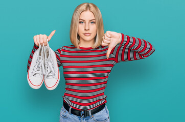 Young blonde woman holding white casual shoes with angry face, negative sign showing dislike with...