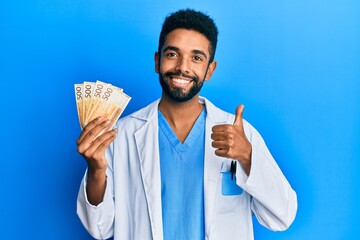 Handsome hispanic man with beard wearing medical uniform holding 500 norwegian krone smiling happy and positive, thumb up doing excellent and approval sign